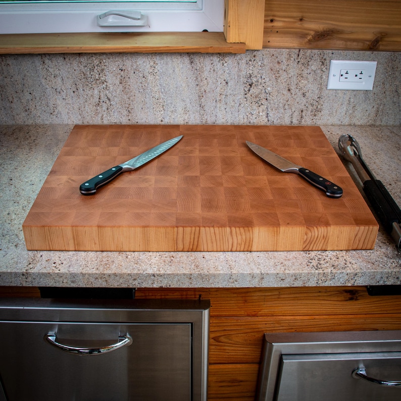 May include: A large, rectangular butcher block cutting board made of light brown wood, with a checkerboard pattern. Two knives with black handles and a pair of tongs rest on the board. The board sits on a speckled countertop.