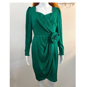 May include: A knee-length emerald green dress featuring a square neckline, long sleeves with gathered shoulders, and a wrap-style skirt. The fabric has a tonal floral pattern and a fabric flower accent at the waist, creating a structured silhouette.