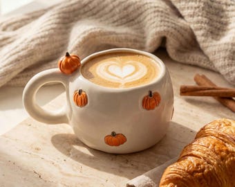 May include: A round white ceramic mug featuring small, raised orange pumpkin decorations on the exterior and a handle. The mug is filled with a coffee beverage topped with heart-shaped latte art, accompanied by a croissant and cinnamon sticks.