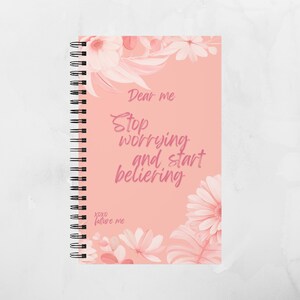 Könnte beinhalten: Ein spiralgebundenes Notizbuch mit korallfarbenem Einband mit floralen Akzenten und dem Text "Dear me, Stop worrying and start believing". Das Notizbuch hat eine schwarze Spiralbindung und ist mit rosa Blumen verziert.