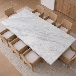 Puede incluir: Vista aérea de un comedor. Una gran mesa rectangular de mármol está rodeada por diez sillas de mimbre con cojines blancos. El conjunto está sobre una alfombra de color neutro, con armarios al fondo.