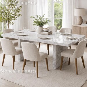 May include: A marble dining table with six upholstered chairs. The table is rectangular with a grey and white veined pattern. The chairs have light beige fabric and wooden legs. The room is bright with natural light and a neutral colour palette.