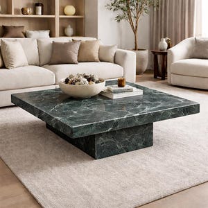 May include: A square coffee table with a dark green marble top and a solid base. The table is in a living room setting with a beige sofa, cushions, and a rug. A decorative bowl and books are on the table.