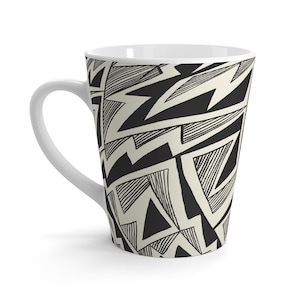 May include: White ceramic latte mug with a black and cream geometric pattern. The design features bold black shapes and lines against a cream background. The mug has a curved handle and a tapered shape.