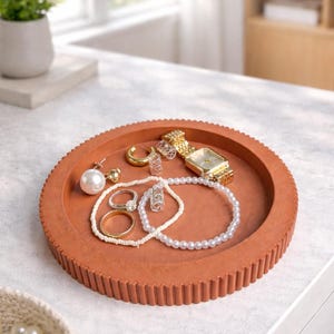 May include: A terracotta-colored, round tray with a textured rim holds various jewelry items. These include a gold watch, rings, earrings, and pearl necklaces and bracelets. The tray sits on a white surface, with a small plant visible in the background.