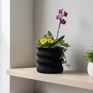 May include: A black, textured, wavy-edged planter holding yellow and purple flowers and greenery, next to a small white pot with a fern. The plants sit on a light wood shelf. Books are visible on a lower shelf.