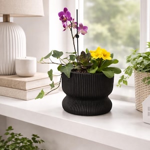 May include: A black, ribbed ceramic planter with a variety of plants, including a purple orchid, yellow flowers, and trailing ivy. The planter sits on a white windowsill, next to a lamp and other decorative items.