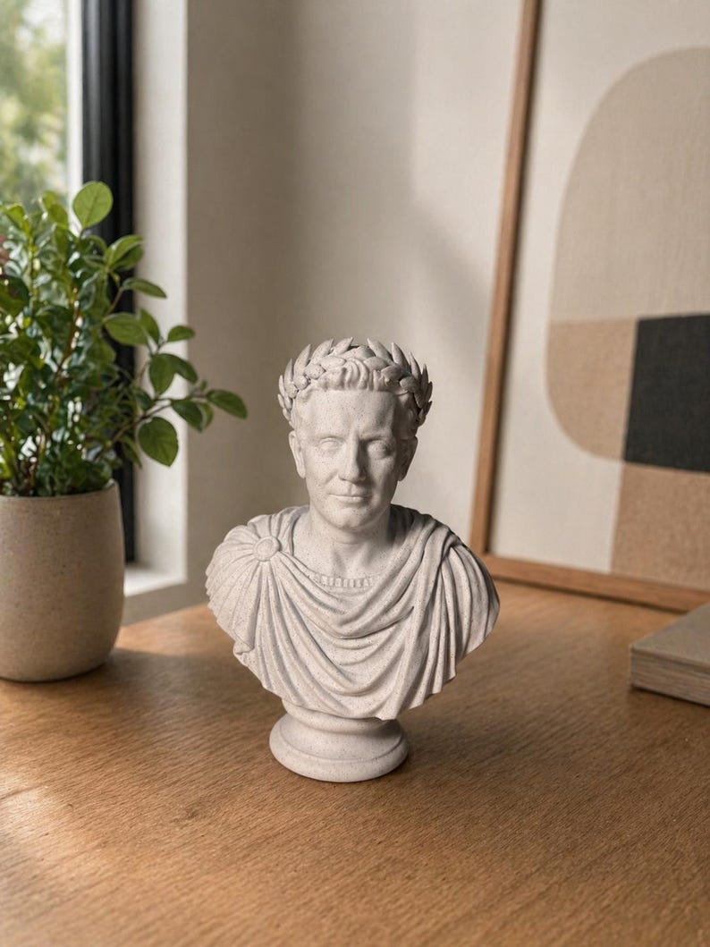 May include: A light gray stone-textured bust of a Roman figure wearing a laurel wreath crown and a draped toga. The sculpture features detailed facial features, a circular brooch on the shoulder, and a pedestal base, capturing a classic aesthetic.