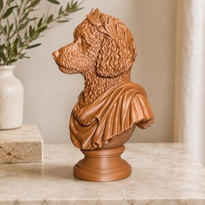 Puede incluir: Un busto escult&oacute;rico de color terracota de un perro de pelo rizado en perfil. El perro lleva una corona de laurel en la cabeza y una prenda drapeada estilo toga sobre los hombros. La escultura est&aacute; montada sobre una base de pedestal circular.