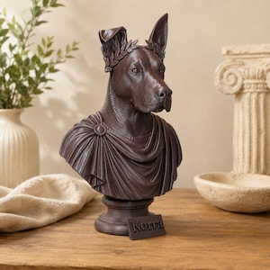 May include: A dark brown bust of a dog wearing a laurel wreath and a draped toga-style garment. The sculpture features a rectangular base with the name Korra embossed on the front. The dog has one ear pointed upward and one ear folded down.