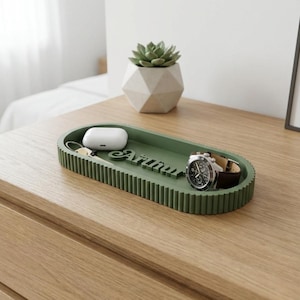 Puede incluir: Una bandeja ovalada verde con borde acanalado contiene un reloj, auriculares inalámbricos y llaves. La bandeja tiene el nombre "Arinnu" recortado. Una pequeña suculenta en una maceta geométrica está en el fondo.