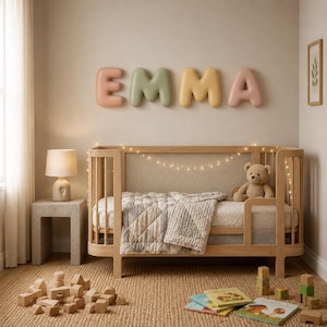 May include: A wooden crib with a light-colored quilt and a teddy bear, featuring wall-mounted, rounded, pastel-colored letters spelling EMMA. A lamp sits on a side table, and wooden building blocks and books are on the floor.