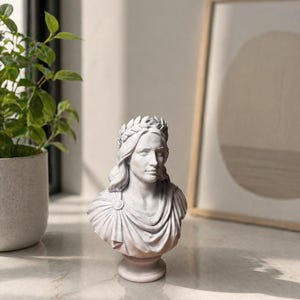 May include: A light gray bust sculpture depicting a figure with long hair adorned with a laurel wreath. The figure wears a draped garment secured with a circular brooch at the shoulder. The sculpture is mounted on a circular pedestal base.