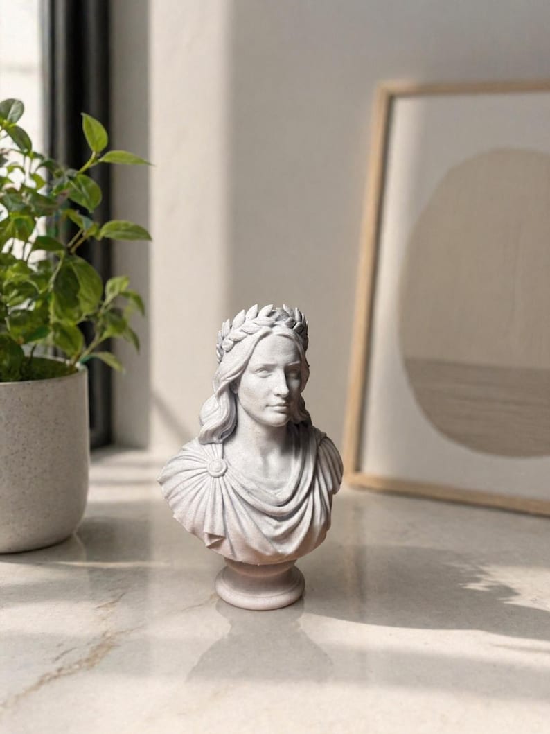 May include: A light gray bust sculpture depicting a figure with long hair adorned with a laurel wreath. The figure wears a draped garment secured with a circular brooch at the shoulder. The sculpture is mounted on a circular pedestal base.