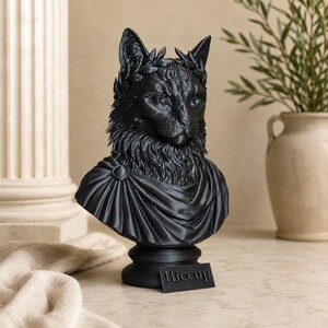 Puede incluir: Un busto negro mate de un gato que lleva una corona de laurel y una prenda drapeada estilo toga. La escultura est&aacute; montada sobre una base circular con una peque&ntilde;a placa rectangular que presenta el texto Hiccup en relieve.