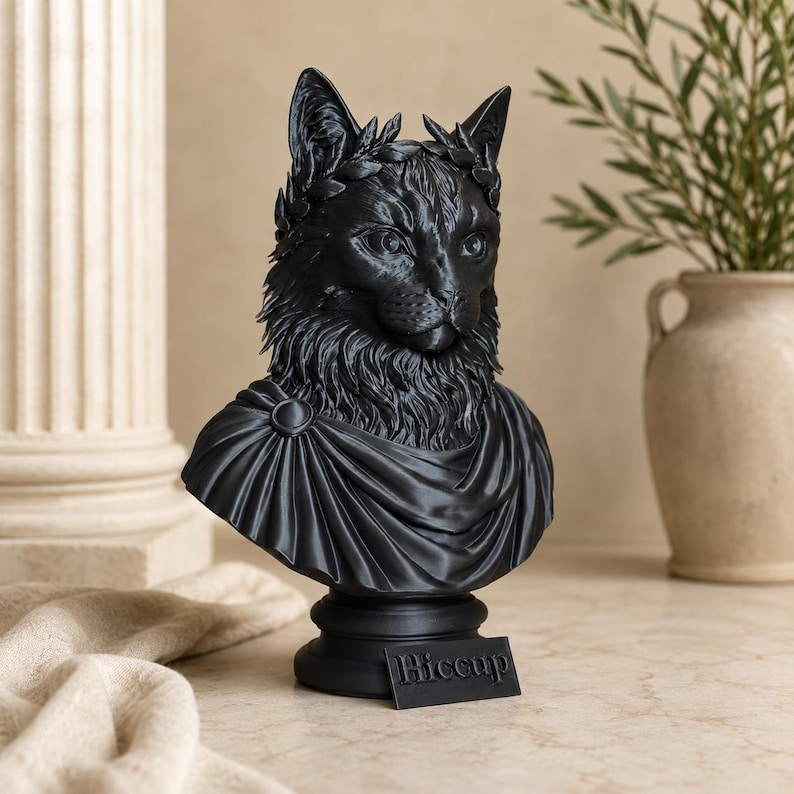 Puede incluir: Un busto negro mate de un gato que lleva una corona de laurel y una prenda drapeada estilo toga. La escultura est&aacute; montada sobre una base circular con una peque&ntilde;a placa rectangular que presenta el texto Hiccup en relieve.