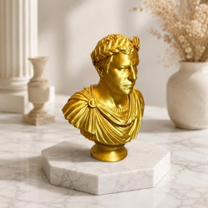 May include: A gold-colored bust of a Roman figure wearing a laurel wreath and a draped toga. The sculpture features detailed facial expressions and fabric folds, resting on a circular base that is mounted on a white marble octagonal pedestal.