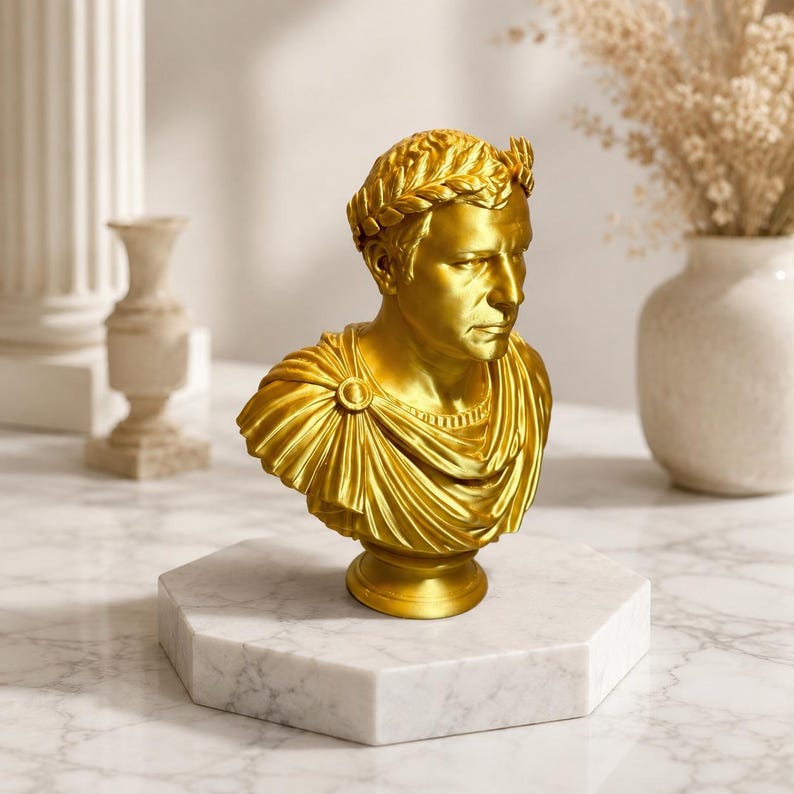 May include: A gold-colored bust of a Roman figure wearing a laurel wreath and a draped toga. The sculpture features detailed facial expressions and fabric folds, resting on a circular base that is mounted on a white marble octagonal pedestal.