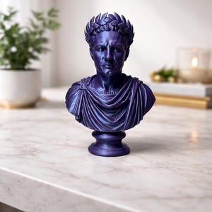 May include: A metallic purple bust of a Roman figure wearing a laurel wreath crown and a draped toga. The sculpture features detailed facial expressions, a circular brooch on the shoulder, and a pedestal base, finished with a smooth, glossy sheen.