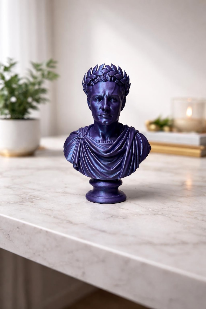 May include: A metallic purple bust of a Roman figure wearing a laurel wreath crown and a draped toga. The sculpture features detailed facial expressions, a circular brooch on the shoulder, and a pedestal base, finished with a smooth, glossy sheen.