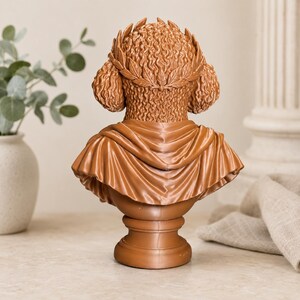 Puede incluir: Una escultura de busto color terracota que muestra la vista posterior de una figura con cabello rizado adornado con una corona de laurel. La figura viste una prenda drapeada sobre los hombros, sobre una base de pedestal circular lisa.