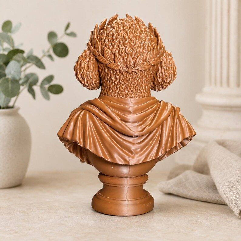 Puede incluir: Una escultura de busto color terracota que muestra la vista posterior de una figura con cabello rizado adornado con una corona de laurel. La figura viste una prenda drapeada sobre los hombros, sobre una base de pedestal circular lisa.