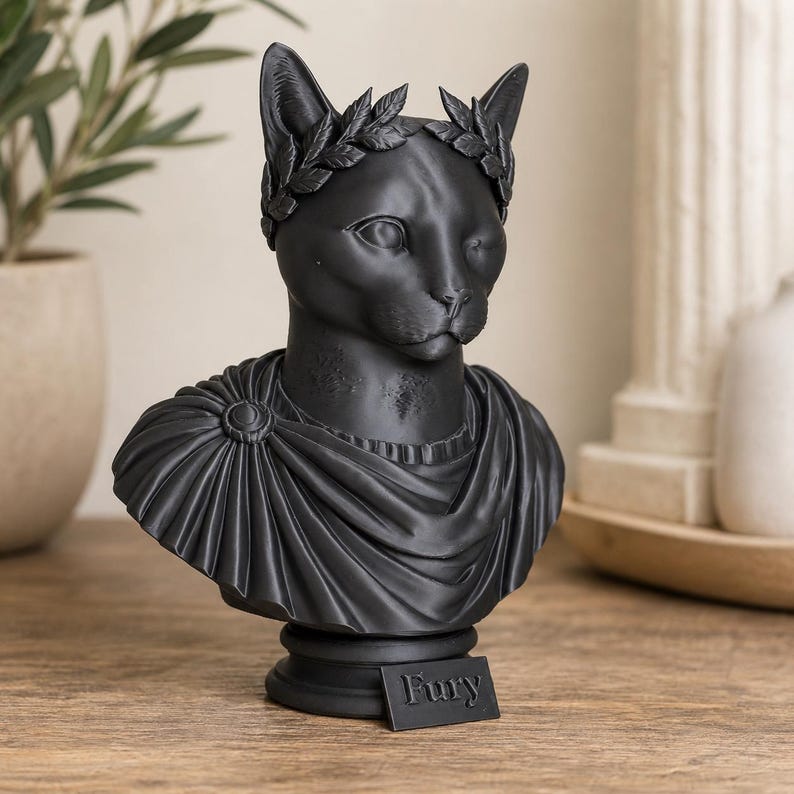 May include: A matte black bust of a cat wearing a laurel wreath crown and a draped toga-style garment. The sculpture is mounted on a circular pedestal base featuring a small rectangular plaque with the word Fury embossed in capital letters.