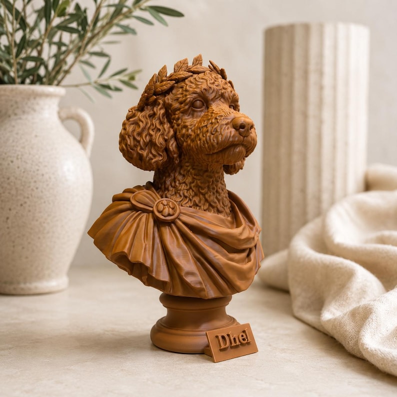 Puede incluir: Un busto de color marr&oacute;n que representa a un perro de pelo rizado con una corona de laurel y una toga drapeada. La escultura incluye una peque&ntilde;a placa rectangular en la base con el texto Dhel grabado en letras de bloque en relieve.