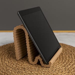 May include: A tan, wavy-shaped tablet stand with a scalloped edge design, holding a black tablet at an angled position. The stand features a continuous, repeating curved structure that provides a stable base for the electronic device.