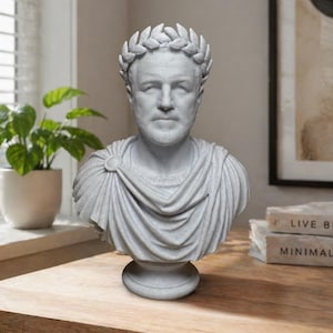 May include: A light gray bust of a Roman figure wearing a laurel wreath crown and a draped toga. The sculpture features detailed facial features, a circular brooch on the shoulder, and a pedestal base, crafted with a textured stone-like finish.