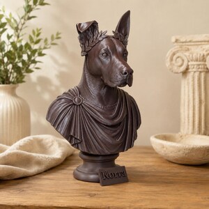 Puede incluir: Un busto de perro de color marr&oacute;n oscuro que lleva una corona de laurel y una prenda drapeada estilo toga. La escultura presenta un broche circular en el hombro y una placa rectangular en la base con el texto "Korra" grabado en relieve.