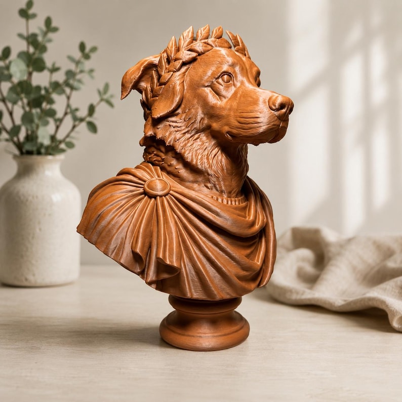 Puede incluir: Un busto de perro color terracota que lleva una corona de laurel en la cabeza y una prenda drapeada estilo toga sujeta con un broche circular en el hombro. La escultura est&aacute; montada sobre una base circular y presenta una textura de pelaje detallada.
