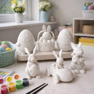 Puede incluir: Colección de decoraciones de Pascua de cerámica blanca, que incluye figuras de conejos, huevos de Pascua y un conejo en un coche. También hay botes de pintura y pinceles, lo que sugiere un tema de manualidades. La escena está sobre una superficie clara.