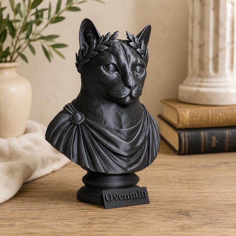 Puede incluir: Un busto negro mate de un gato que lleva una corona de laurel y una prenda drapeada estilo toga. La escultura est&aacute; montada sobre una base circular con una peque&ntilde;a placa rectangular fijada en la parte frontal con el texto en relieve Ovemmit.