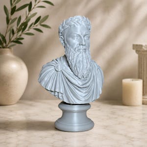 May include: A light gray bust sculpture of a bearded man wearing a laurel wreath on his head and a draped toga fastened with a circular brooch at the shoulder. The figure is mounted on a smooth, circular pedestal base with a matte finish.