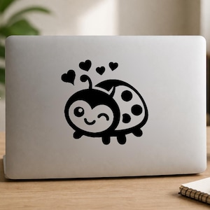 May include: A black vinyl decal sticker applied to the lid of a silver laptop. The design features a cartoon ladybird with a winking face, spots on its back, and four small floating hearts positioned above its head in a simple graphic style.