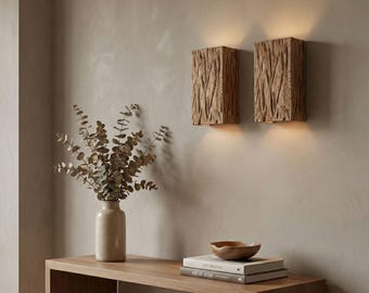 May include: Two rectangular wooden wall sconces with a textured, bark-like appearance are mounted on a neutral-colored wall. The sconces emit a warm, inviting light. A wooden console table holds a vase with dried foliage, books, and a wooden bowl, creating a cozy interior design.