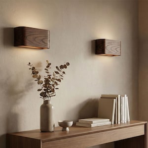 Op de afbeelding: Twee houten wandkandelaars met een rechthoekig ontwerp zijn aan een beige muur bevestigd. De wandkandelaars geven een warm licht af. Een houten console eronder bevat een vaas met gedroogde takken, een kleine kom en boeken.