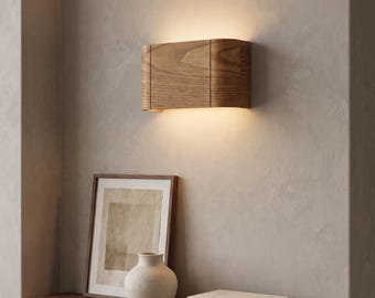 Puede incluir: Un aplique de pared rectangular fabricado en madera con un patrón de veta visible. El accesorio presenta un diseño curvo que emite un brillo cálido y suave desde los bordes superior e inferior, iluminando la pared por encima y por debajo.