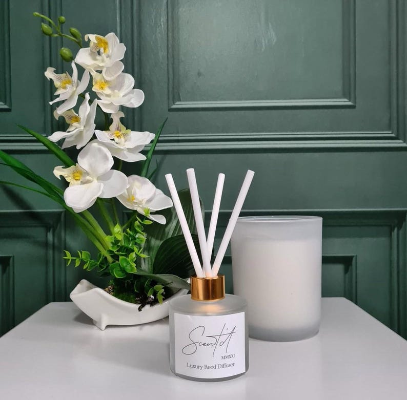 Peut inclure: Un diffuseur de parfum &agrave; roseaux blanc avec un col dor&eacute; et des roseaux blancs, &agrave; c&ocirc;t&eacute; d'une bougie blanche et d'une composition florale d'orchid&eacute;es blanches. Le diffuseur porte le texte "Scent it". Fond vert fonc&eacute;.