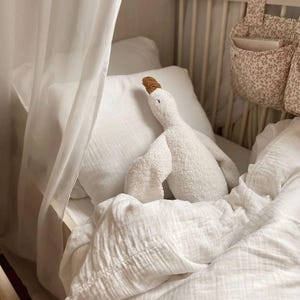 Puede incluir: Un juguete de peluche de ganso blanco con pico y patas marrones descansa sobre una almohada y ropa de cama blancas. La ropa de cama es en capas y texturizada. Una bolsa de almacenamiento con estampado floral cuelga del lado de la cuna.