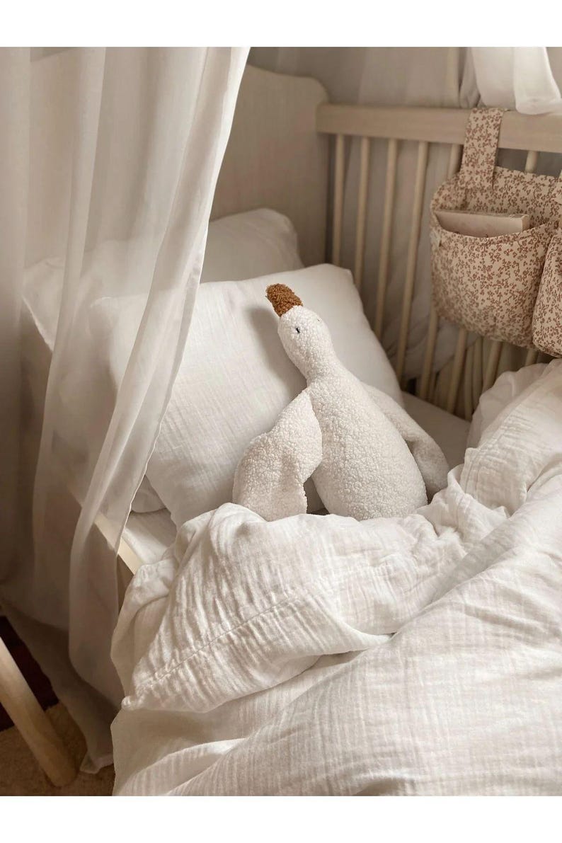 Puede incluir: Un juguete de peluche de ganso blanco con pico y patas marrones descansa sobre una almohada y ropa de cama blancas. La ropa de cama es en capas y texturizada. Una bolsa de almacenamiento con estampado floral cuelga del lado de la cuna.