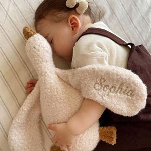 May include: A plush goose toy with a textured cream-coloured fleece exterior, featuring a brown beak and feet. The name Sophia is embroidered in cursive script on one of the goose's wings. The toy has a soft, stuffed body and long, flexible neck.