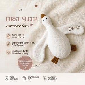 May include: A white plush goose toy with brown feet and a brown beak, featuring the name Olivia embroidered on one wing. The text FIRST SLEEP companion is visible alongside icons and descriptions detailing 100% cotton muslin fabric.