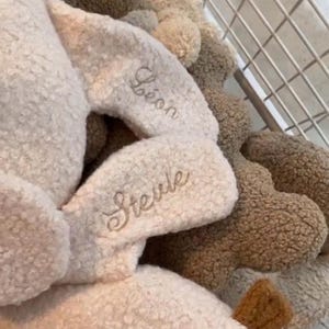 May include: A collection of plush toys made from cream and brown textured sherpa fabric. The toys feature embroidered names including L&eacute;on and Stevie in a cursive script. The items are arranged inside a white wire basket with a grid pattern.