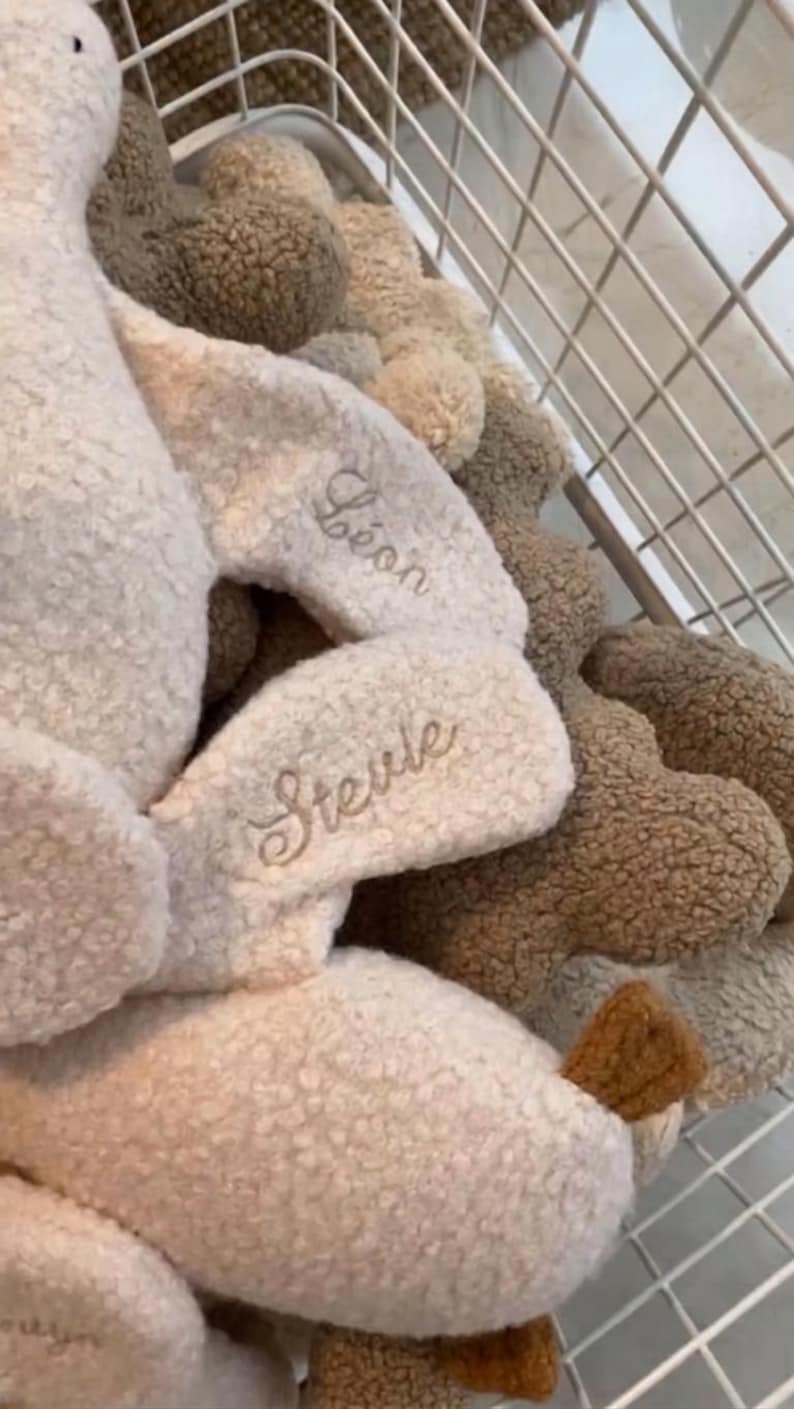 May include: A collection of plush toys made from cream and brown textured sherpa fabric. The toys feature embroidered names including L&eacute;on and Stevie in a cursive script. The items are arranged inside a white wire basket with a grid pattern.