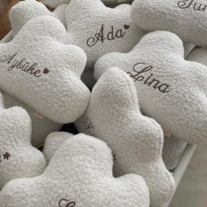 May include: A collection of white, cloud-shaped plush cushions made of textured sherpa fabric. Each cushion features embroidered names in cursive script, including Deniz, Yekta, Tur, Ada, Aybüke, Lina, and Ege, with some names accented by a small heart.