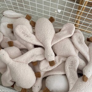 May include: A collection of plush goose toys made from cream-coloured textured fabric. Each toy features brown fabric beaks and feet, with various names embroidered in cursive script on the wings, including Atlas, Alperen, Bade, and Vera.