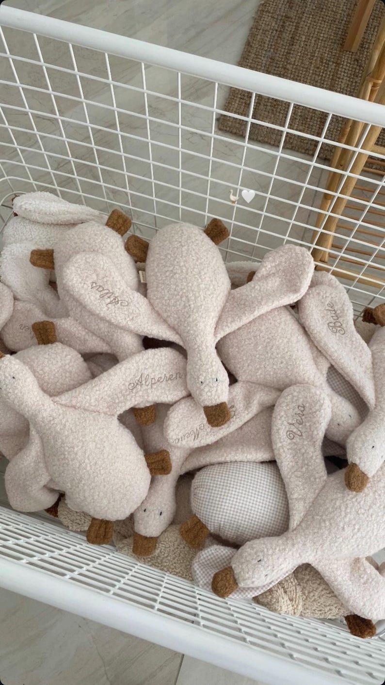 May include: A collection of plush goose toys made from cream-coloured textured fabric. Each toy features brown fabric beaks and feet, with various names embroidered in cursive script on the wings, including Atlas, Alperen, Bade, and Vera.