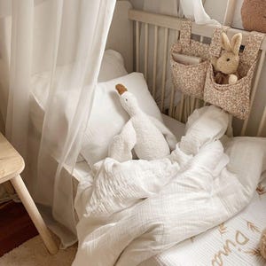 Puede incluir: Una escena de guarder&iacute;a con una cuna blanca, ropa de cama blanca y suave, y un peluche de ganso blanco. Un organizador con estampado floral contiene un conejito de peluche. La cuna est&aacute; cubierta con una cortina blanca transparente.